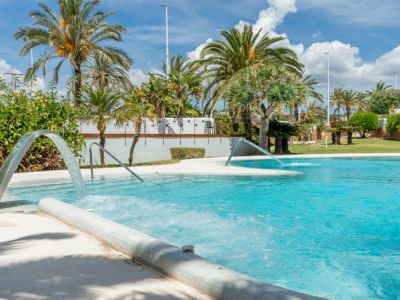 Amplia piscina comunitaria con cascadas de agua y zonas de burbujas, rodeada de exuberante vegetación y palmeras bajo un cielo azul.