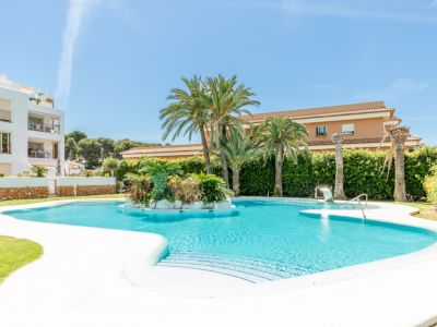 Amplia piscina comunitaria de forma irregular con isla central de palmeras y vegetación exuberante, rodeada de césped y edificios residenciales bajo un cielo despejado.