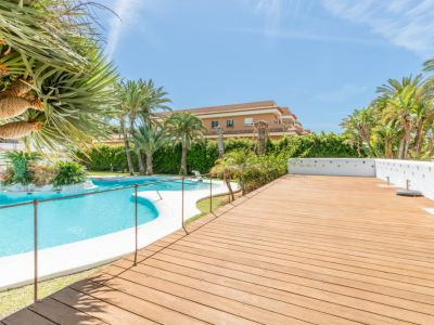 Amplia zona comunitaria con una gran piscina curva y una isla ajardinada central. La espaciosa tarima de madera adyacente está rodeada de palmeras y exuberante vegetación mediterránea.