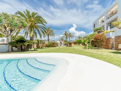 Amplia zona de piscina comunitaria con una gran alberca de escalones de mosaico azul, rodeada de césped verde y palmeras mediterráneas bajo un cielo claro.