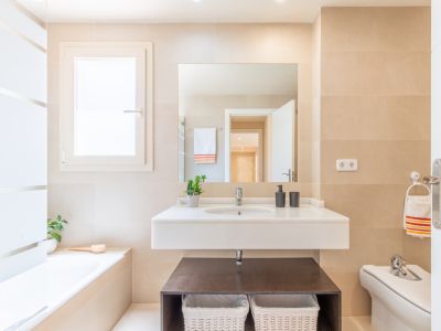 Baño completo con lavabo suspendido, encimera blanca y espejo amplio. Destacan los azulejos claros, ventana con privacidad y bañera integrada.