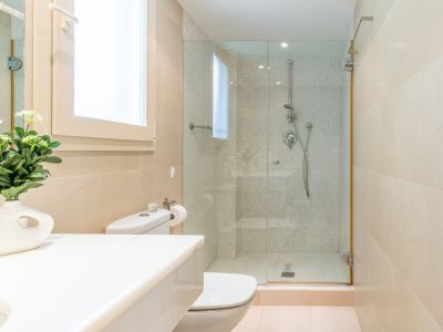 Baño moderno con inodoro, lavabo de encimera blanca y ventana. Ducha acristalada con revestimiento de mosaico y azulejos beige, complementado con luz natural.