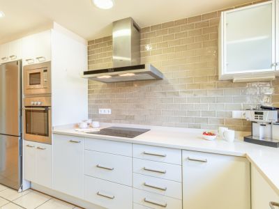 Cocina moderna con mobiliario blanco, electrodomésticos integrados de acero inoxidable y una encimera clara. Destaca el salpicadero de azulejos tipo ladrillo beige y la placa de inducción.