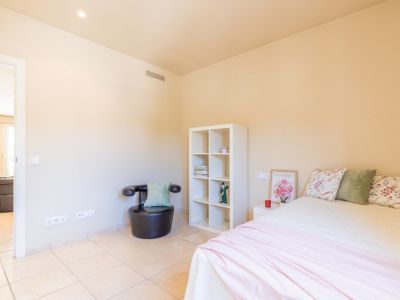Dormitorio luminoso con cama doble, suelo de baldosas claras, paredes en tono crema y detalles decorativos. Incluye estantería blanca, sillón y acceso a otra estancia.