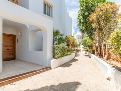 Entrada principal con puerta de madera moderna y fachada blanca arqueada. Un sendero ajardinado con vegetación mediterránea conduce a través del complejo.