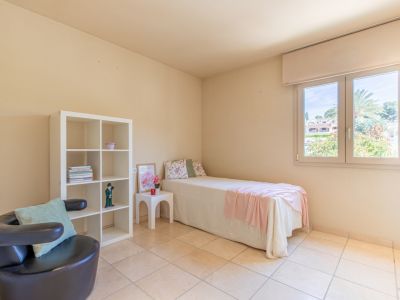 Luminoso dormitorio secundario con cama individual, estantería blanca y sillón oscuro. La ventana ofrece vistas a la vegetación exterior y abundante luz natural sobre el suelo de baldosas claras.