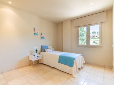 Luminoso dormitorio secundario con cama individual vestida en tonos azules y blancos, suelo de baldosas claras y ventana con vistas a la vegetación y viviendas. Iluminación empotrada.