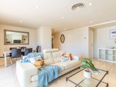 Salón-comedor de diseño abierto con sofá de piel claro, mesa de centro de cristal y suelo de baldosas claras. Iluminación empotrada y ventilación de techo.