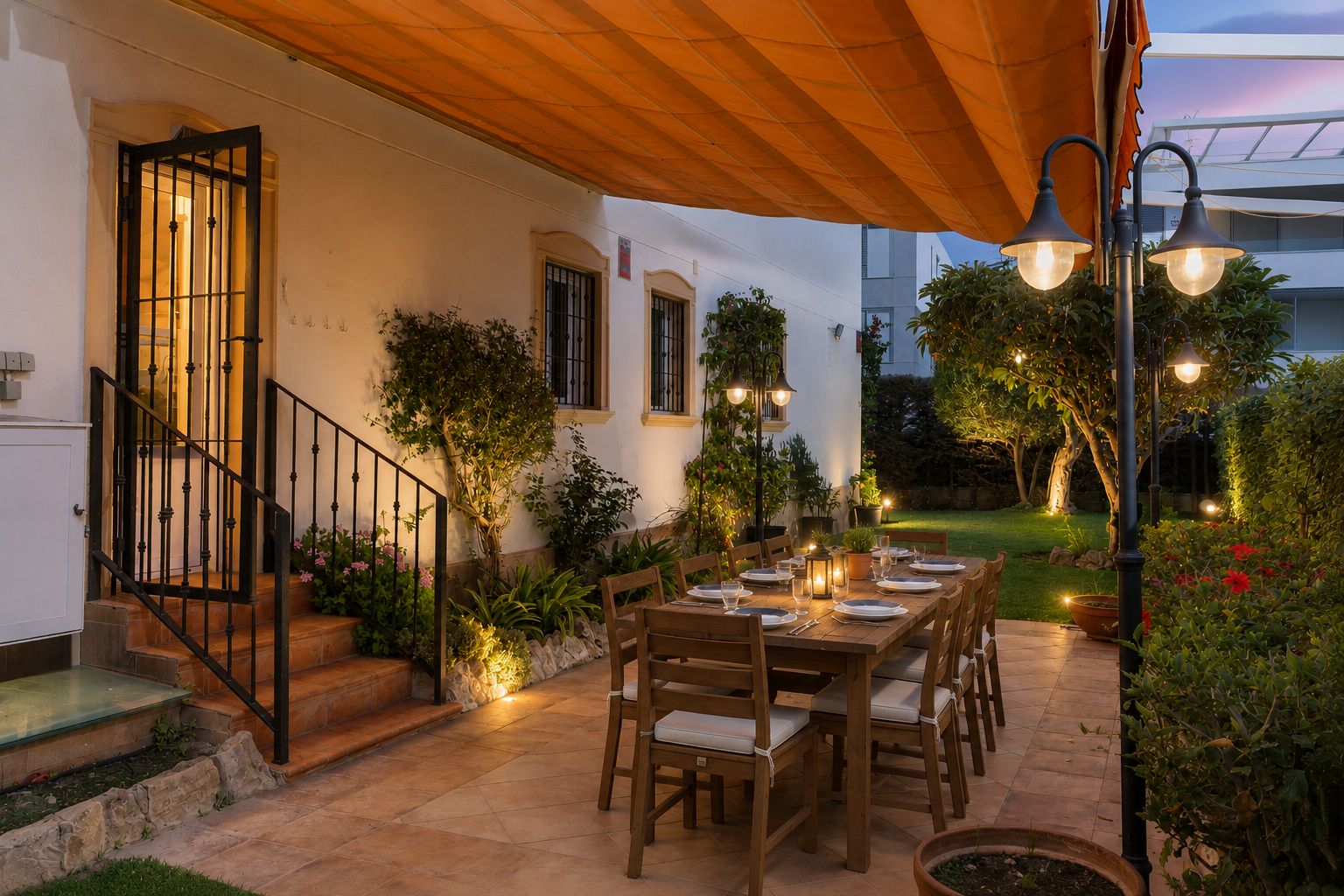 Acogedor comedor exterior en patio embaldosado, con gran mesa de madera preparada bajo toldo naranja retráctil y jardín privado iluminado.
