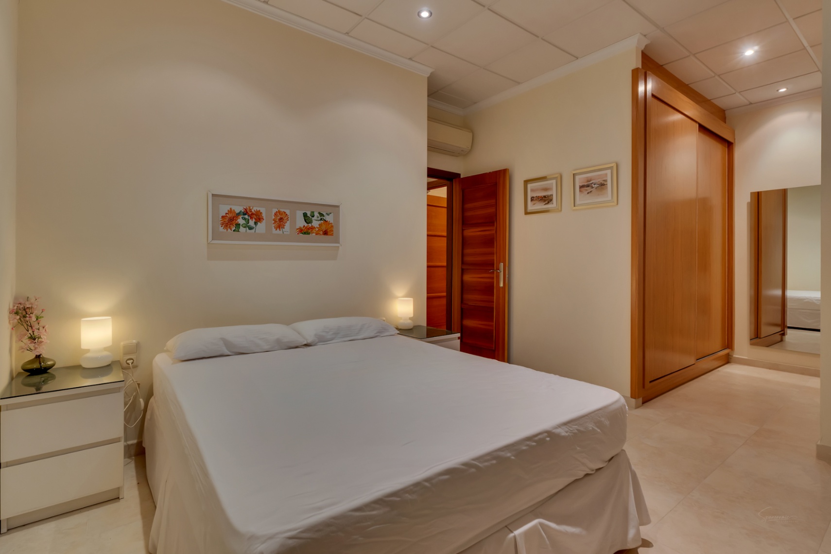 Acogedor dormitorio con cama doble, mesitas de noche modernas, amplio armario empotrado de madera y suelo de baldosas claras, complementado con aire acondicionado.