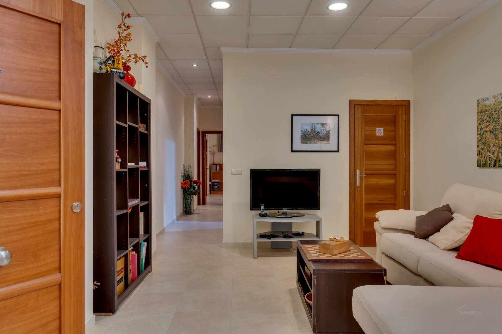 Acogedor salón con sofá, TV y mesa de centro con ajedrez. Destacan las puertas de madera clara, estantería y el pasillo que se extiende al fondo.