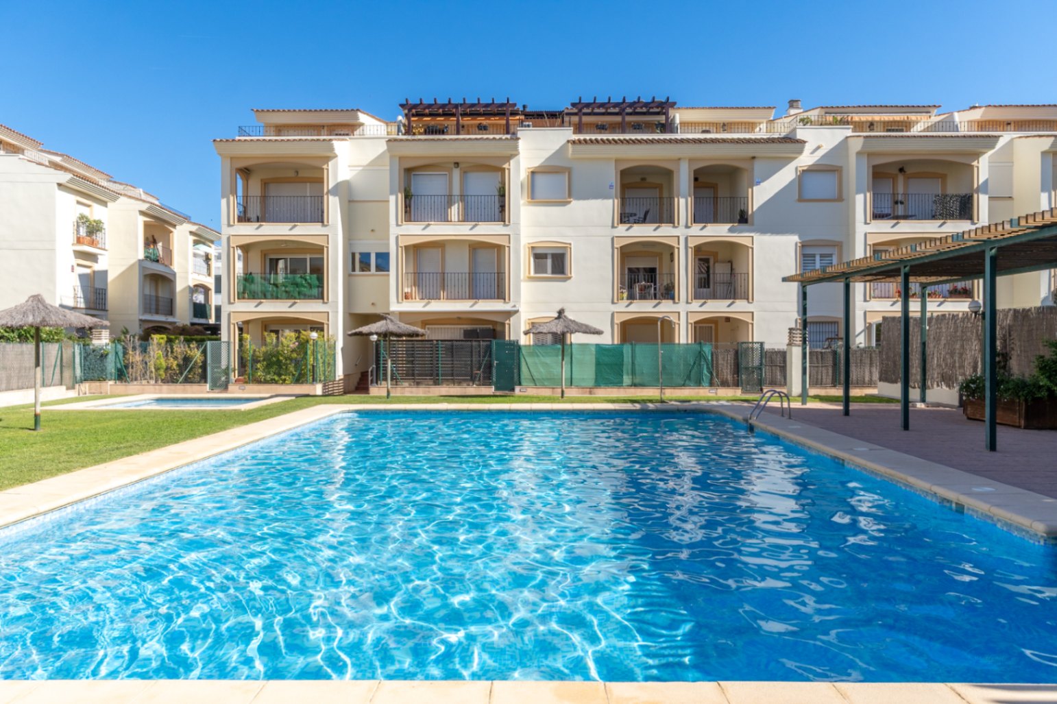 Amplia piscina comunitaria de agua salada, rodeada de césped verde y edificios de apartamentos color crema con balcones privados.