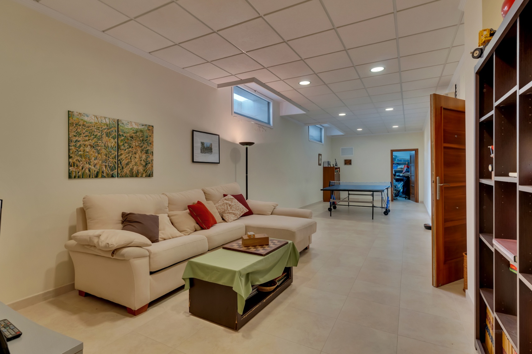 Amplia sala de estar en planta inferior con sofá en L, mesa de centro, mesa de ping-pong y ventanas altas, ideal como zona de juegos o segundo salón.
