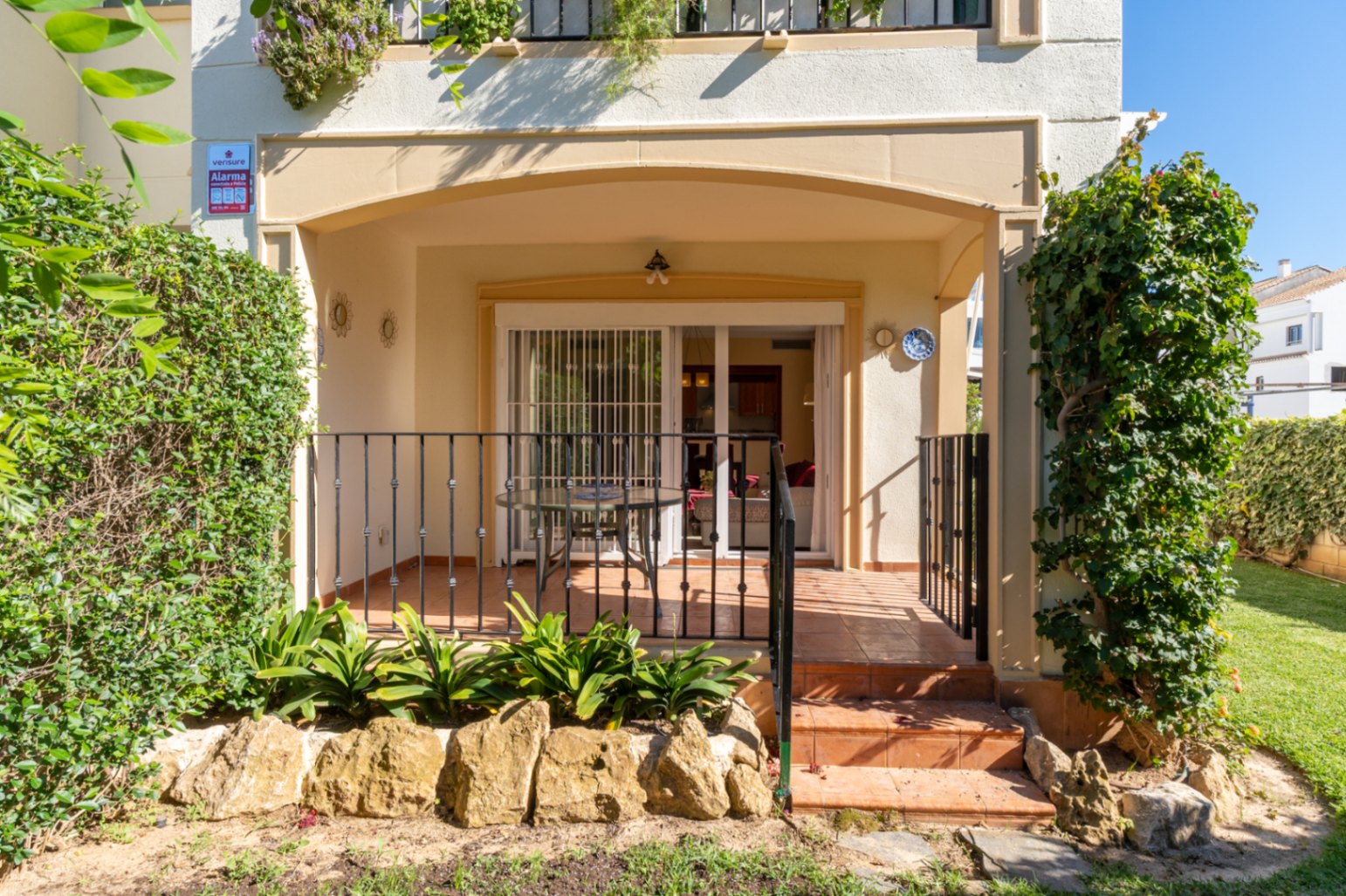 Amplia terraza cubierta con suelo de baldosas de terracota, barandilla de metal negro y acceso directo al jardín privado con setos y césped.