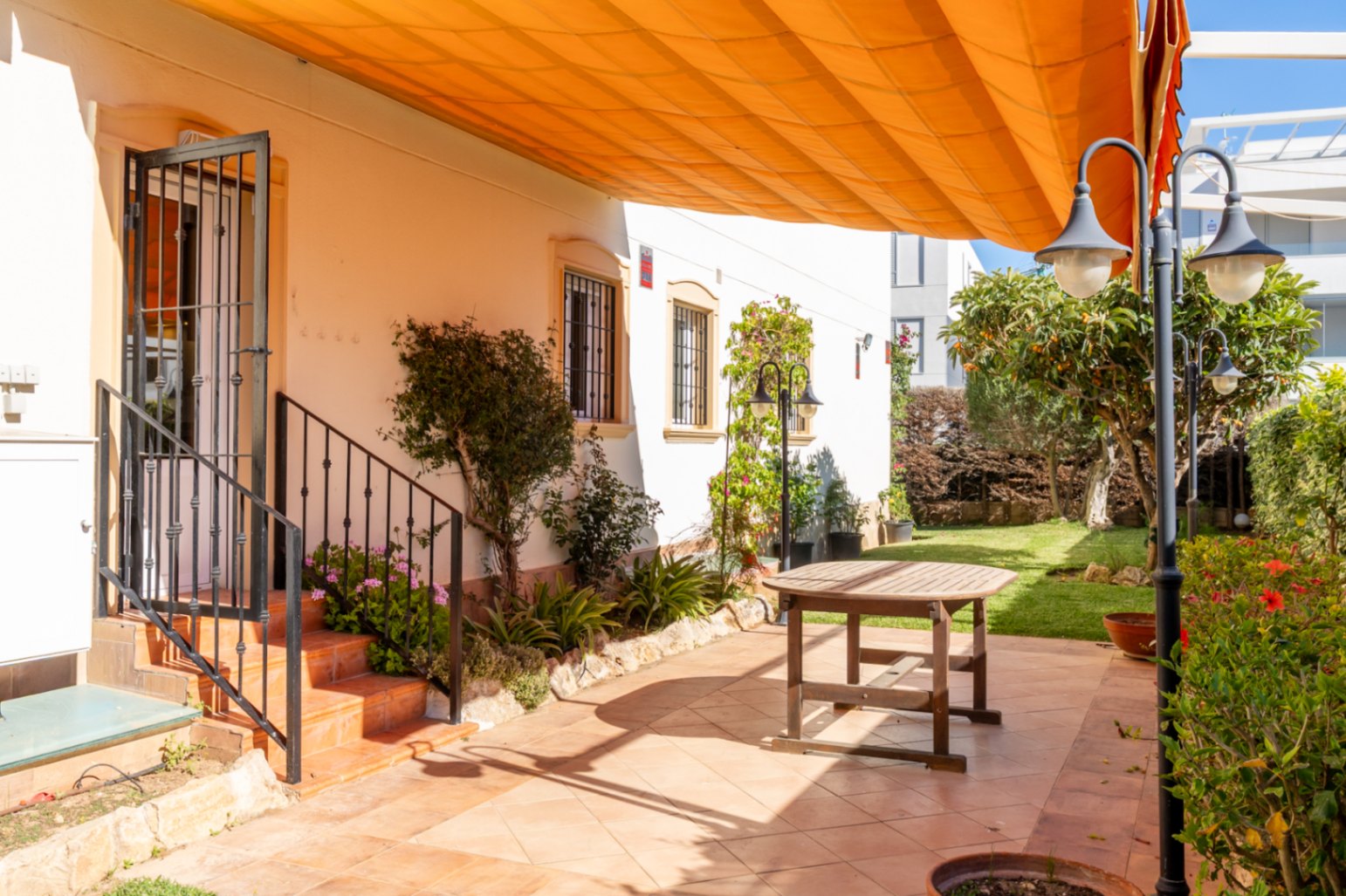 Amplio patio exterior con suelo de baldosas de terracota, mesa de madera y toldo naranja retráctil. Acceso a la vivienda y jardín con césped y vegetación.