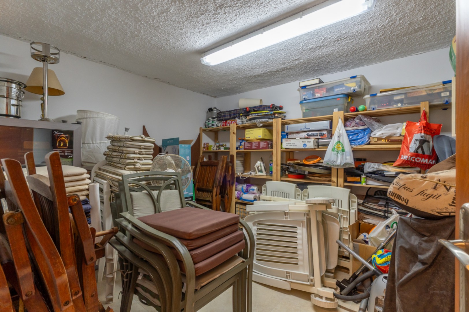 Amplio trastero con estanterías de madera repletas de objetos domésticos, muebles de jardín apilados y una luminaria fluorescente en el techo.