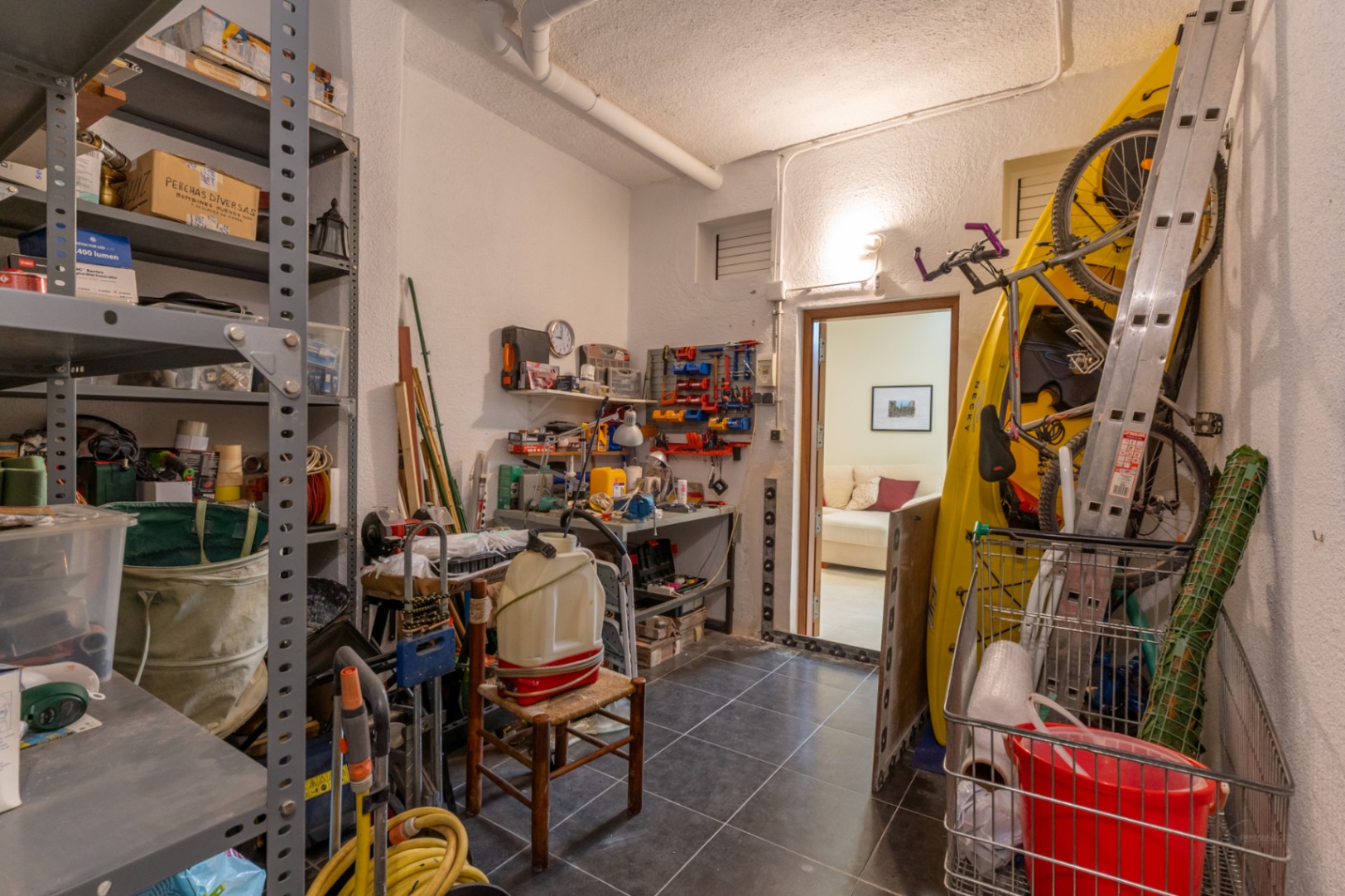 Amplio trastero con suelo de baldosas oscuras, estanterías metálicas repletas, un banco de trabajo y almacenamiento para kayak y bicicleta, con acceso a una sala interior.