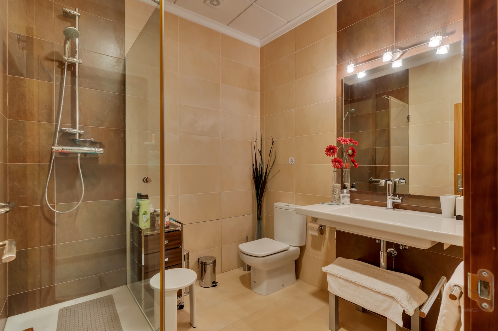 Baño completo con ducha acristalada, azulejos en tonos beige y marrón, lavabo suspendido y espejo con iluminación moderna.