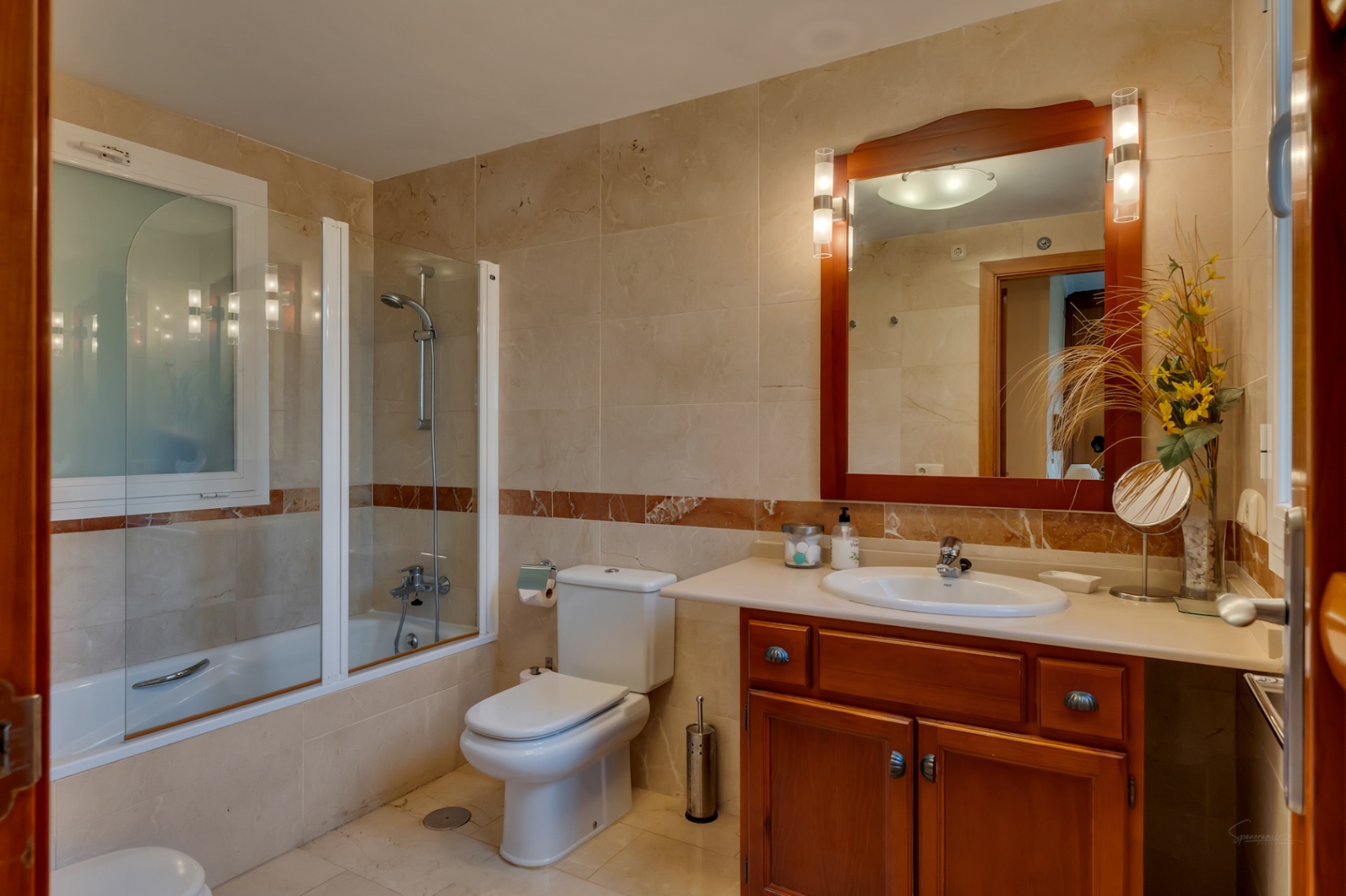 Baño con azulejos beige, banda decorativa de mármol y bañera con mampara de cristal. Incluye tocador de madera con lavabo y espejo iluminado, además de una ventana esmerilada.