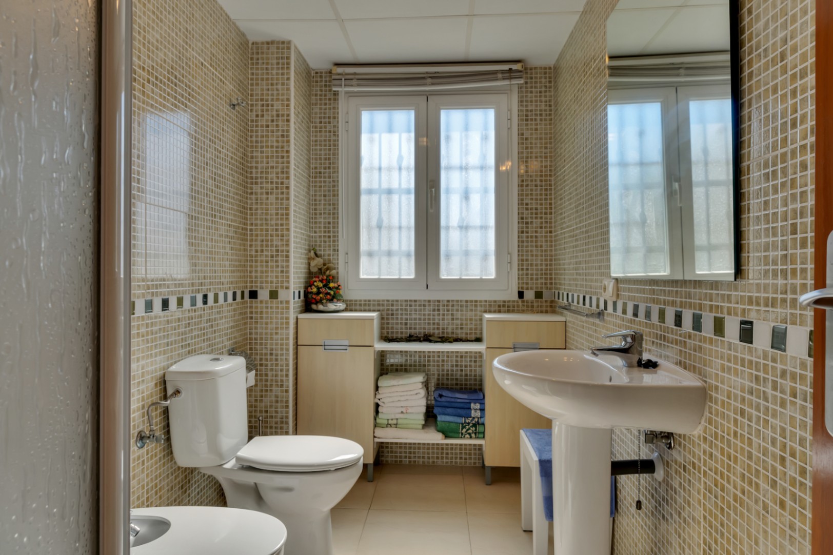 Baño con paredes de mosaico claro y cenefa decorativa, lavabo de pedestal, inodoro y ventana que aporta luz natural. Muebles bajos de madera clara bajo la ventana para almacenaje.