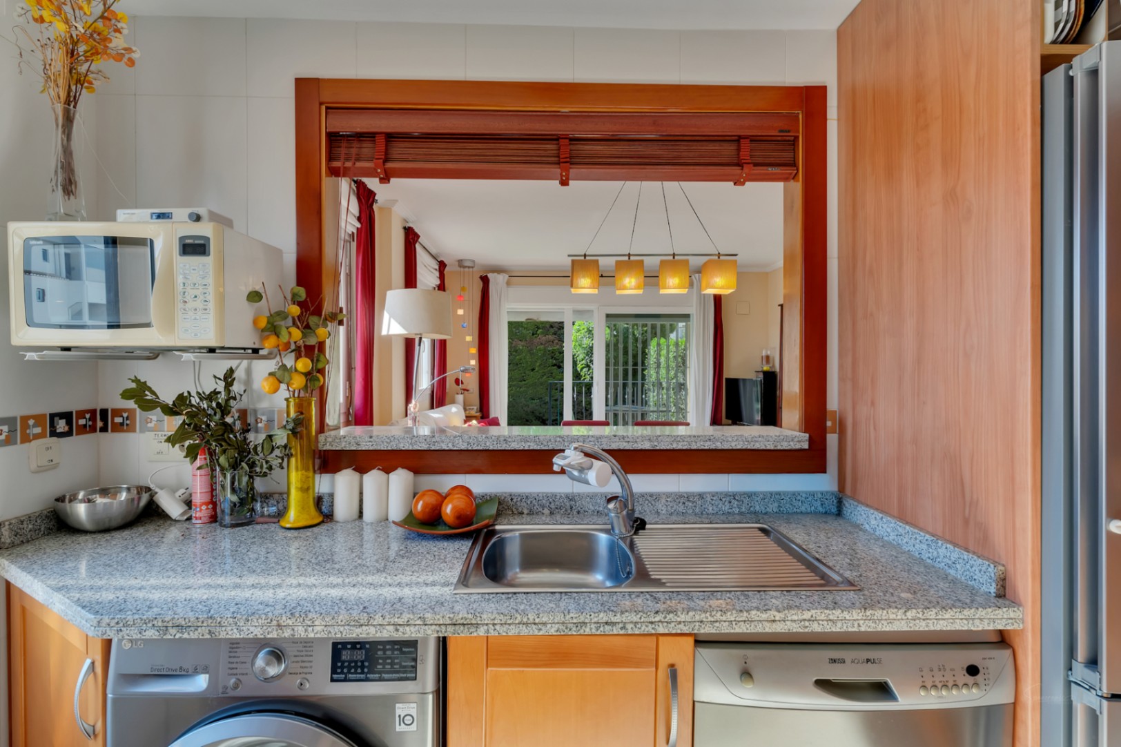 Cocina funcional con encimera de granito, fregadero de acero, lavadora y lavavajillas integrados. Una ventana pasaplatos de madera conecta con el salón-comedor.