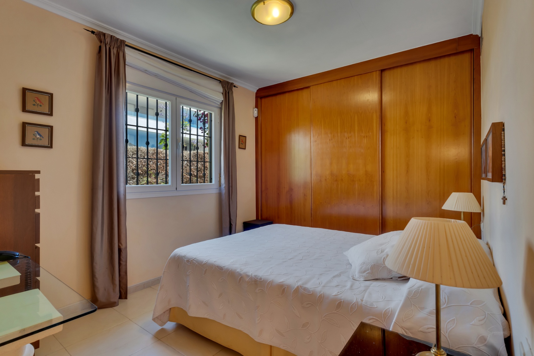 Dormitorio con cama doble, armario empotrado de madera y ventana con rejas que da a vegetación exterior.