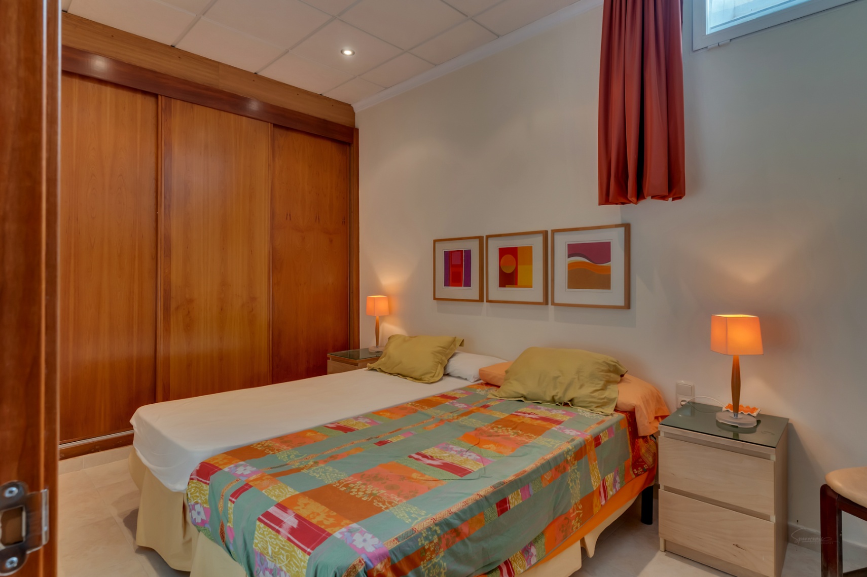 Dormitorio con dos camas individuales, un amplio armario empotrado de madera con puertas correderas y dos mesitas de noche con lámparas cálidas. Arte abstracto en la pared blanca.