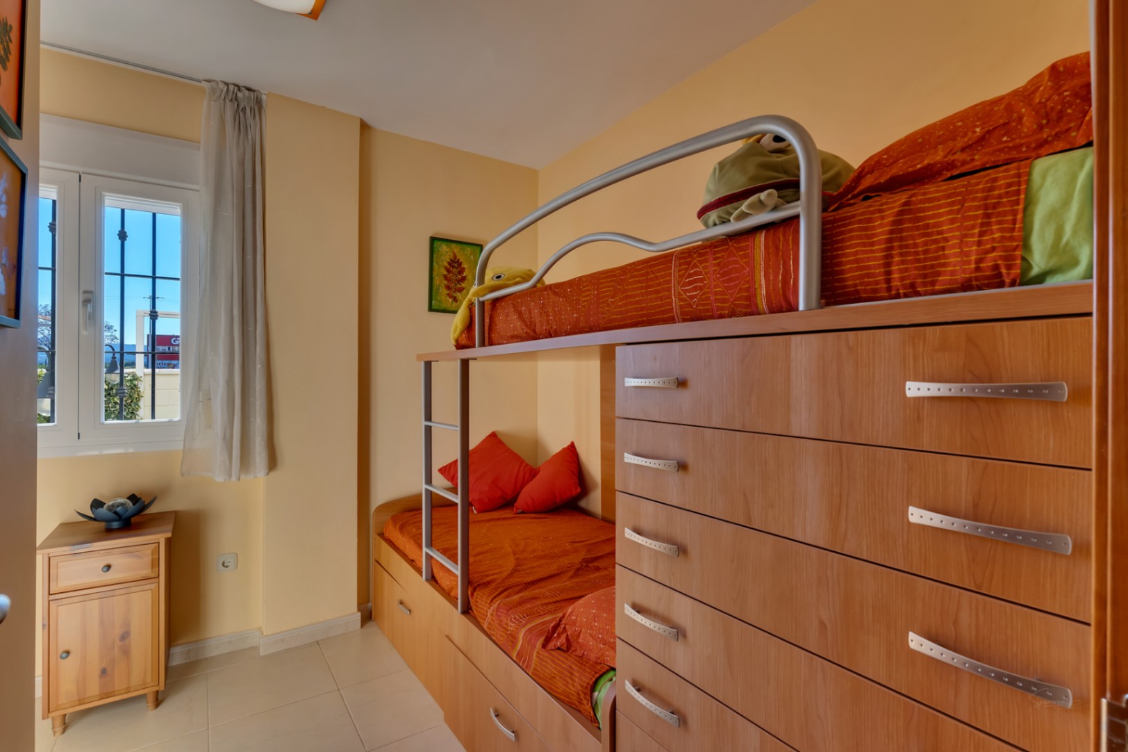 Dormitorio infantil con literas integradas en mueble de madera clara, ofreciendo soluciones de almacenaje. La habitación recibe luz natural a través de una ventana con rejas y cortina translúcida.