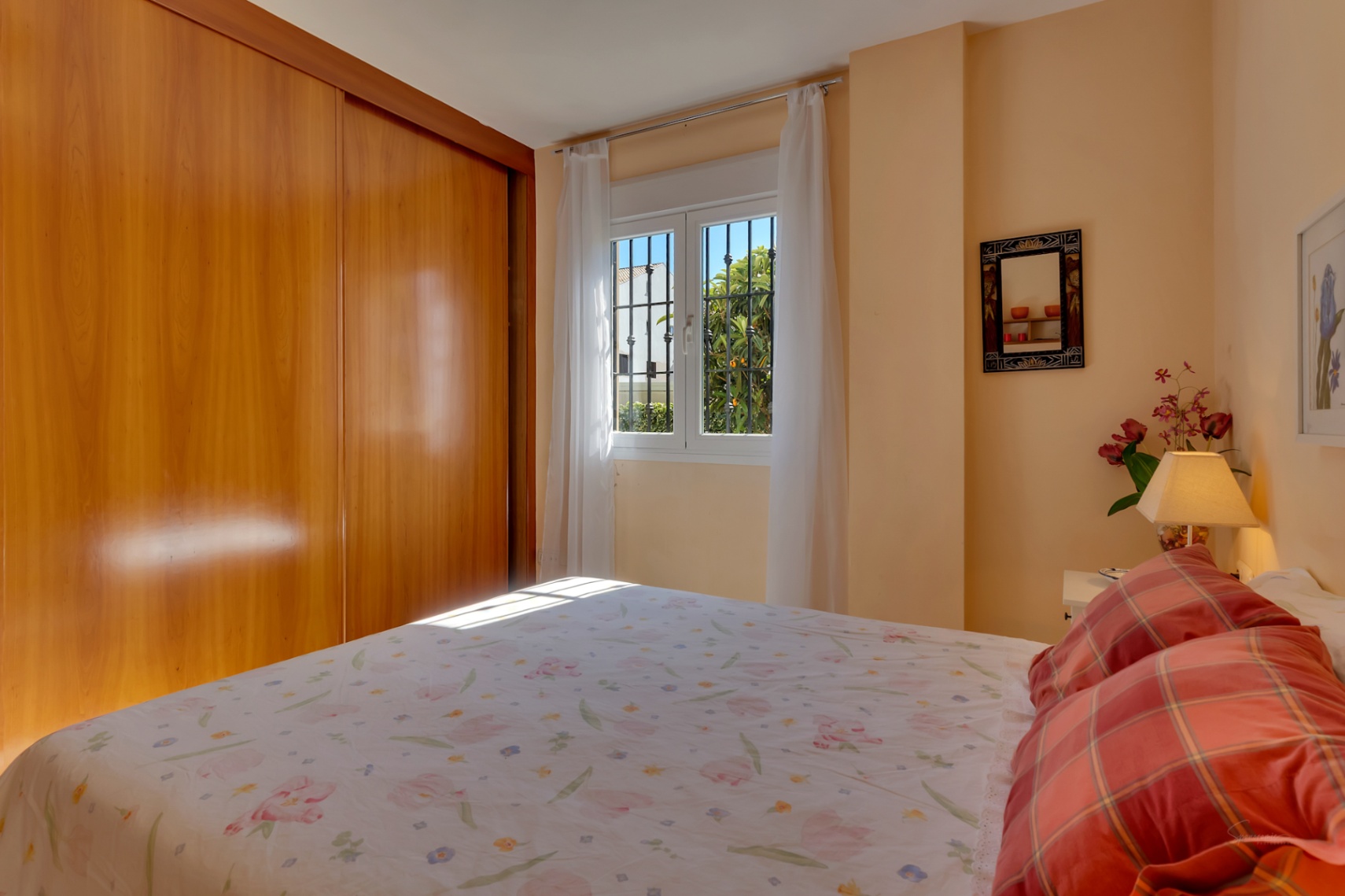 Dormitorio principal con amplio armario empotrado de madera clara, cama doble y ventana con rejas que ofrece luz natural y vistas a la vegetación.