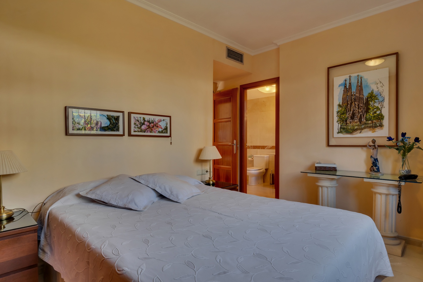 Dormitorio principal con cama doble, mesitas de noche de madera y acceso directo a un baño privado. Destacan cuadros decorativos y una consola de cristal con pilares clásicos.