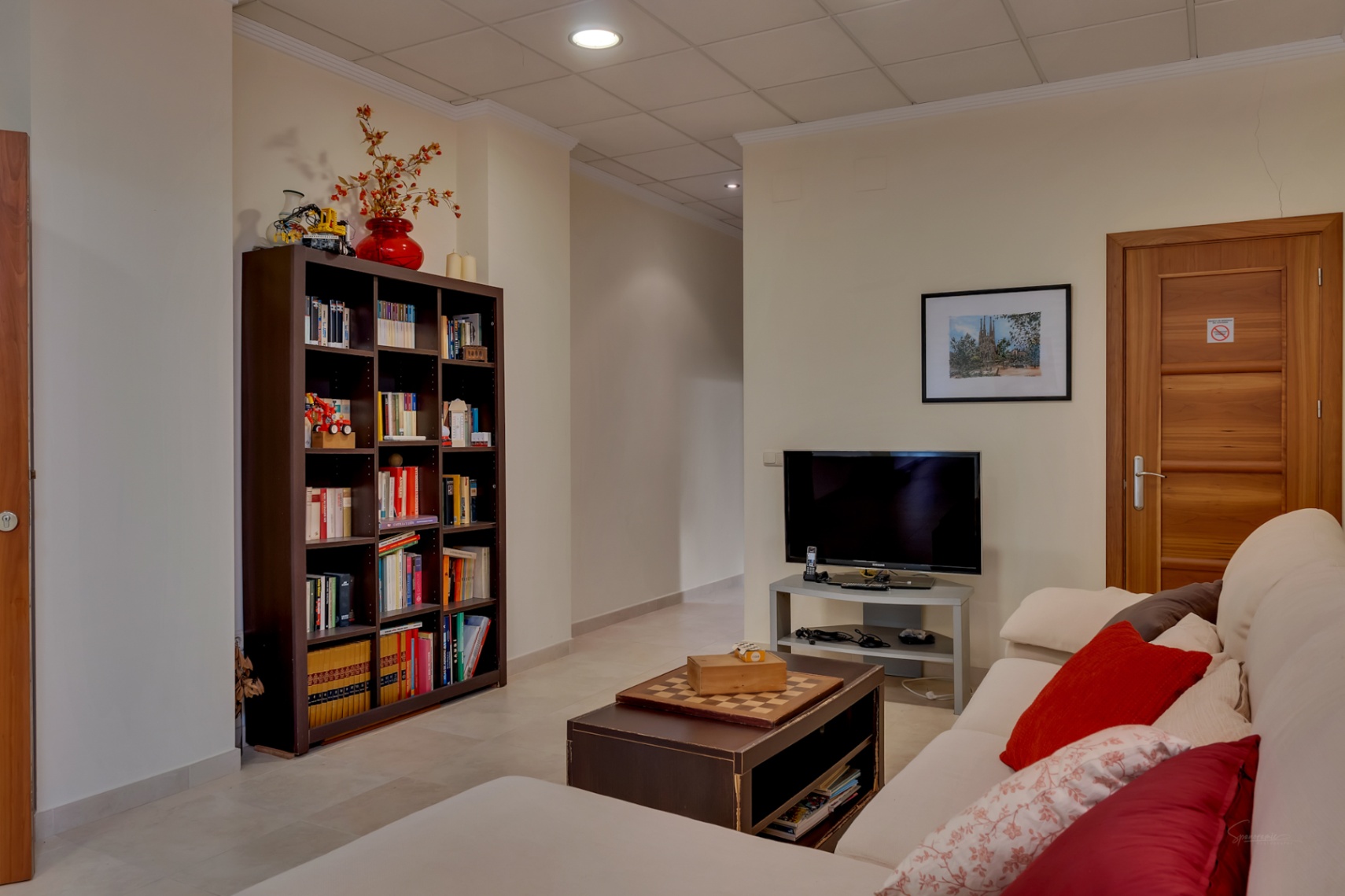 Este versátil salón secundario cuenta con un cómodo sofá, mesa de centro con ajedrez, y una gran estantería llena de libros. Incluye televisión y suelo de baldosas claras.