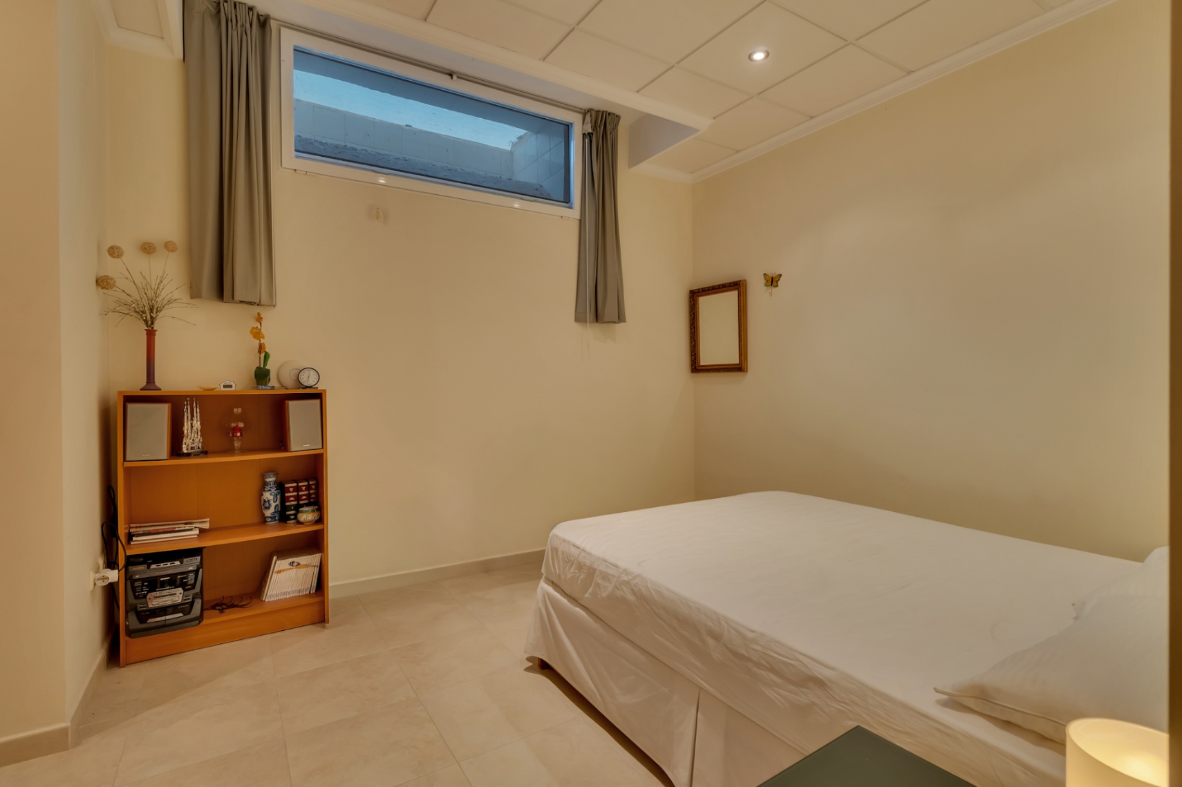 Habitación secundaria con cama individual, suelo de baldosas claras y paredes crema. Incluye una estantería de madera, un sistema de música y una ventana alta con cortinas grises.