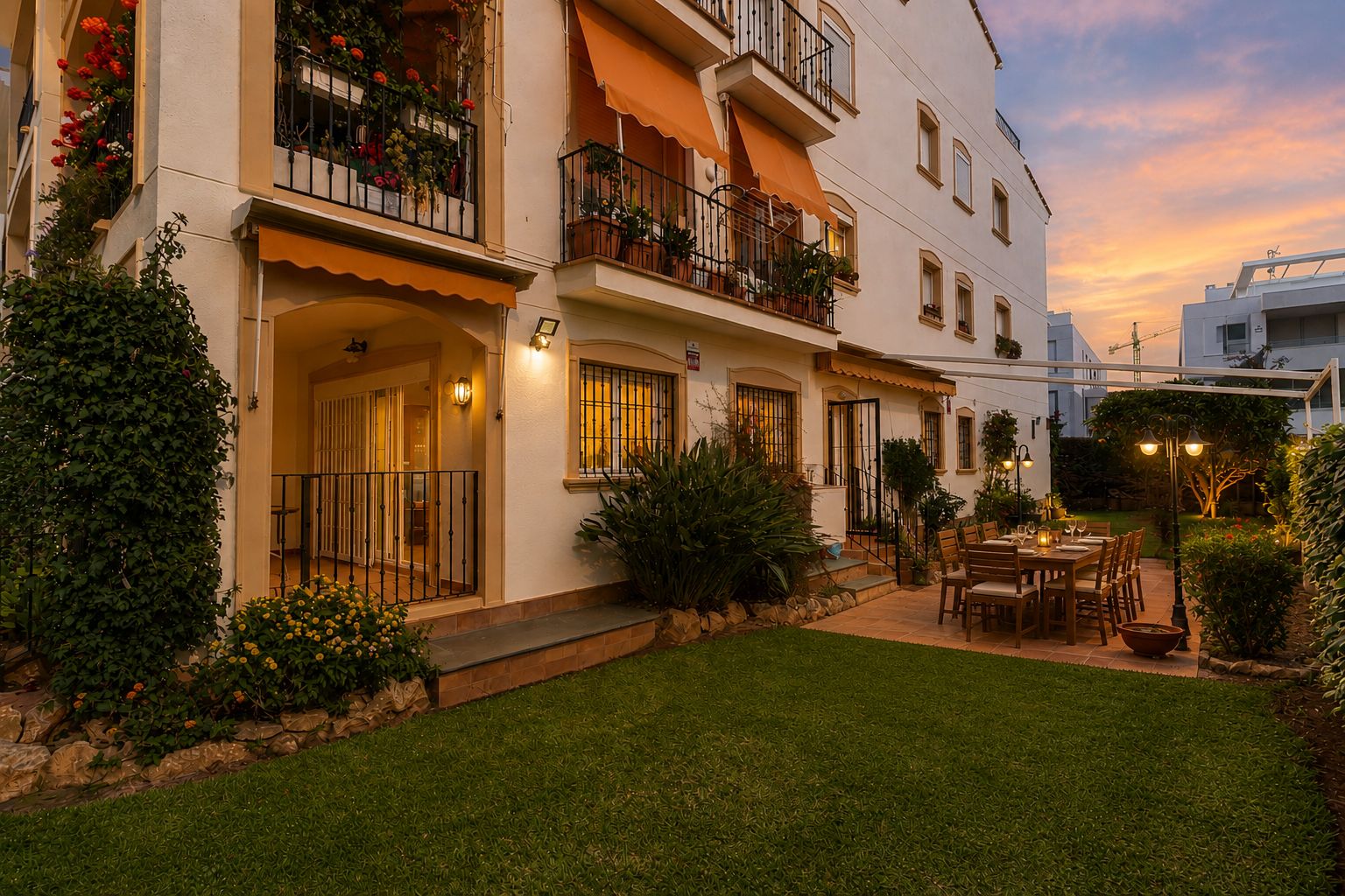 Jardín privado con césped y patio embaldosado, ideal para cenas al aire libre. Destaca la mesa de comedor de madera, la iluminación ambiental y la exuberante vegetación al atardecer.