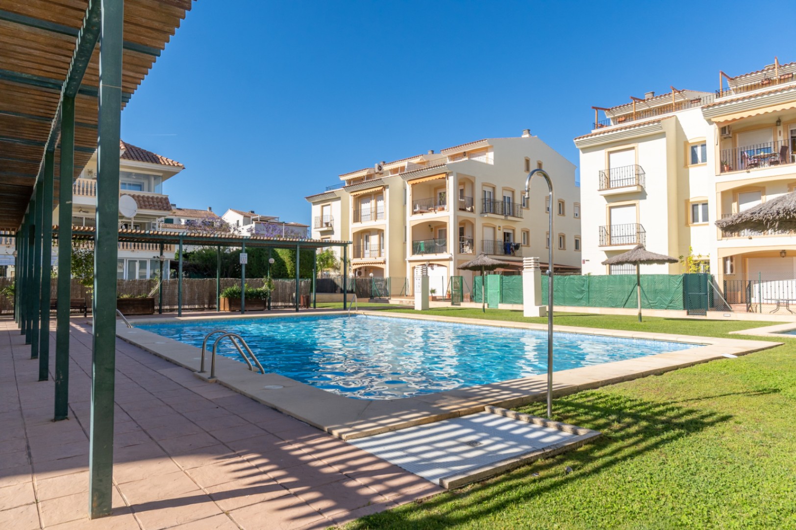 Piscina comunitaria de agua salada con azulejos azules, césped verde y edificios residenciales en el fondo. Pérgola de madera y ducha exterior.