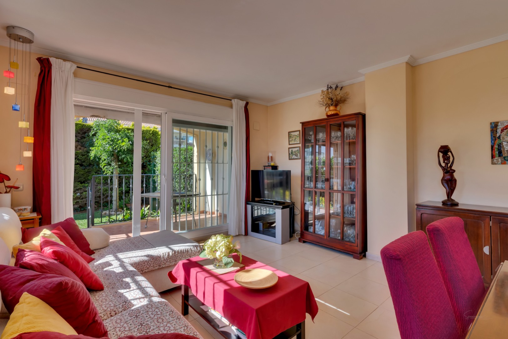 Sala de estar luminosa con sofá en L, cojines rojos y amarillos. Acceso directo a terraza y jardín privado a través de puertas correderas, con mueble vitrina de madera y lámpara colgante.
