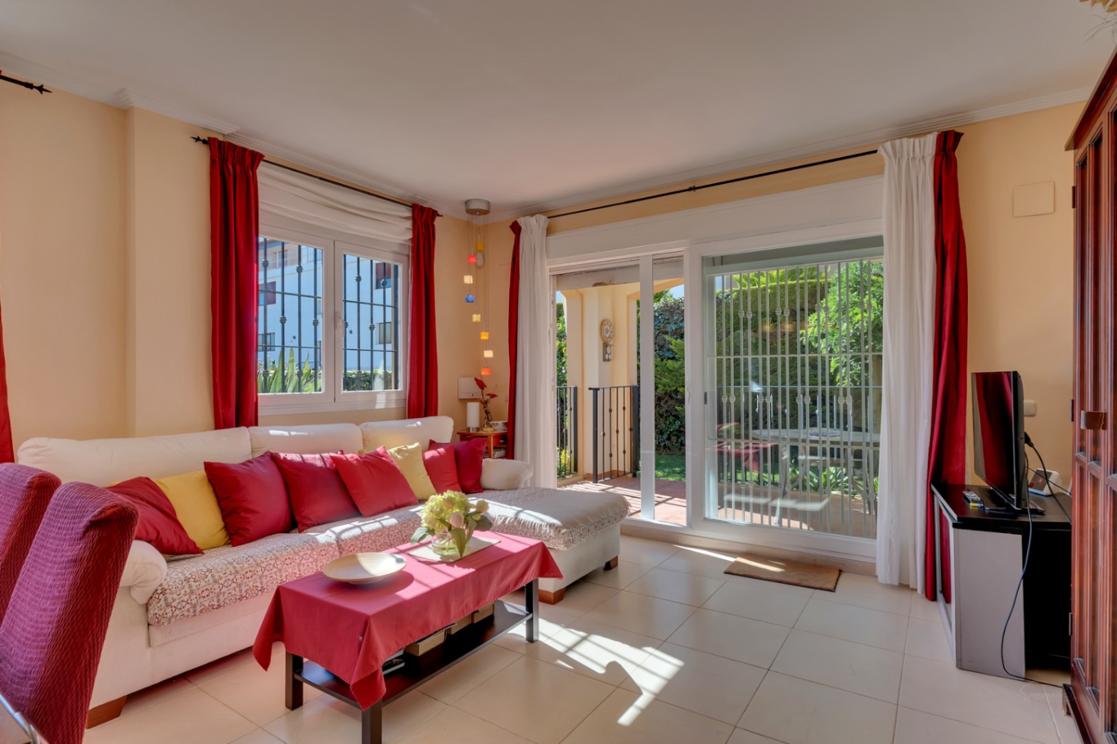 Salón luminoso con sofá blanco, cojines vibrantes, suelo de baldosas y acceso directo al jardín privado a través de puertas correderas.