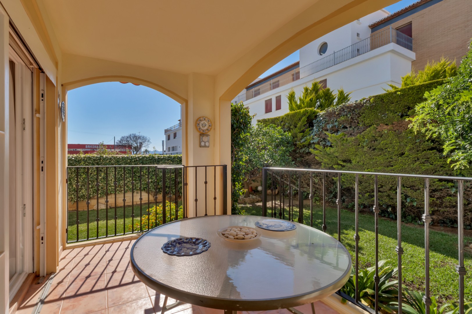Terraza cubierta con mesa de cristal y suelo de terracota, con vistas directas a un jardín privado con césped y setos, y edificios distantes bajo luz natural.