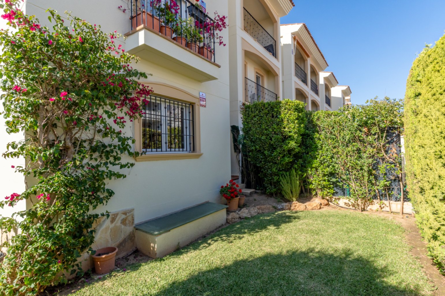 Vista del jardín privado con césped verde, buganvilla en flor en la fachada y setos. Incluye una ventana con rejas y un banco exterior.