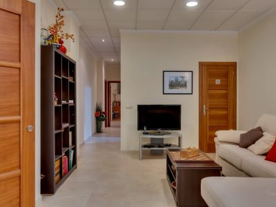 Acogedor salón con sofá, TV y mesa de centro con ajedrez. Destacan las puertas de madera clara, estantería y el pasillo que se extiende al fondo.