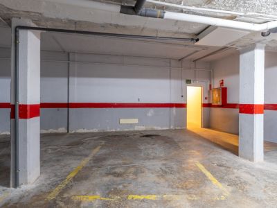 Amplia plaza de garaje subterráneo con suelo de hormigón, marcas amarillas y tuberías expuestas. Incluye puerta de salida iluminada y equipo contra incendios.