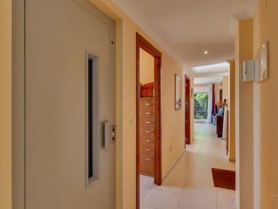 Amplio pasillo con paredes crema y suelo de baldosas blancas, destacando un ascensor integrado y múltiples puertas de madera. Al fondo, se vislumbra un área luminosa con acceso al jardín.
