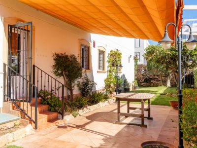 Amplio patio exterior con suelo de baldosas de terracota, mesa de madera y toldo naranja retráctil. Acceso a la vivienda y jardín con césped y vegetación.