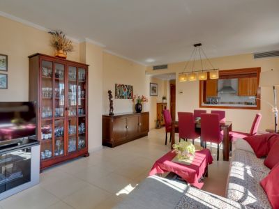 Amplio salón-comedor con sofá claro y detalles rojos, mesa de comedor de madera con sillas a juego y vitrina. Una ventana pasaplatos conecta con la cocina, y la luz natural ilumina el espacio.