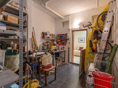 Amplio trastero con suelo de baldosas oscuras, estanterías metálicas repletas, un banco de trabajo y almacenamiento para kayak y bicicleta, con acceso a una sala interior.