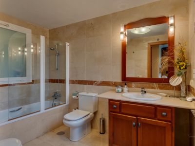 Baño con azulejos beige, banda decorativa de mármol y bañera con mampara de cristal. Incluye tocador de madera con lavabo y espejo iluminado, además de una ventana esmerilada.