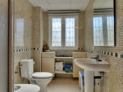 Baño con paredes de mosaico claro y cenefa decorativa, lavabo de pedestal, inodoro y ventana que aporta luz natural. Muebles bajos de madera clara bajo la ventana para almacenaje.