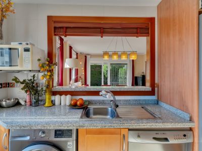 Cocina funcional con encimera de granito, fregadero de acero, lavadora y lavavajillas integrados. Una ventana pasaplatos de madera conecta con el salón-comedor.