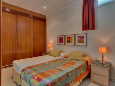 Dormitorio con dos camas individuales, un amplio armario empotrado de madera con puertas correderas y dos mesitas de noche con lámparas cálidas. Arte abstracto en la pared blanca.