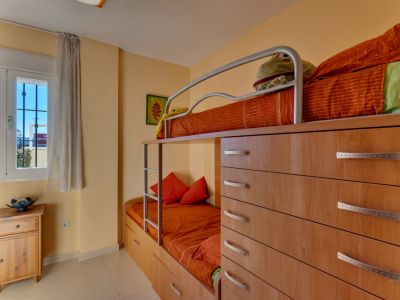 Dormitorio infantil con literas integradas en mueble de madera clara, ofreciendo soluciones de almacenaje. La habitación recibe luz natural a través de una ventana con rejas y cortina translúcida.