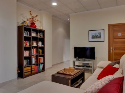 Este versátil salón secundario cuenta con un cómodo sofá, mesa de centro con ajedrez, y una gran estantería llena de libros. Incluye televisión y suelo de baldosas claras.